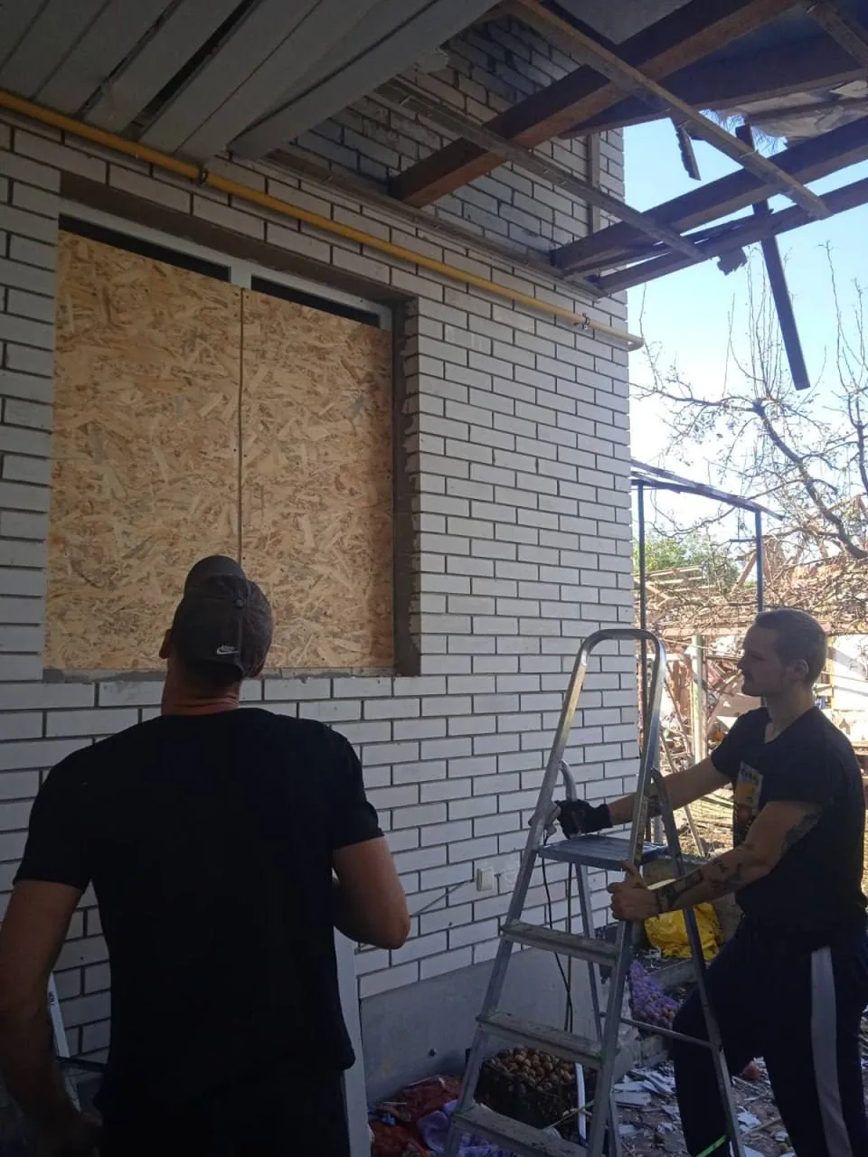 Двоє учасників програми біля закритих плитами вікон пошкодженого будинку